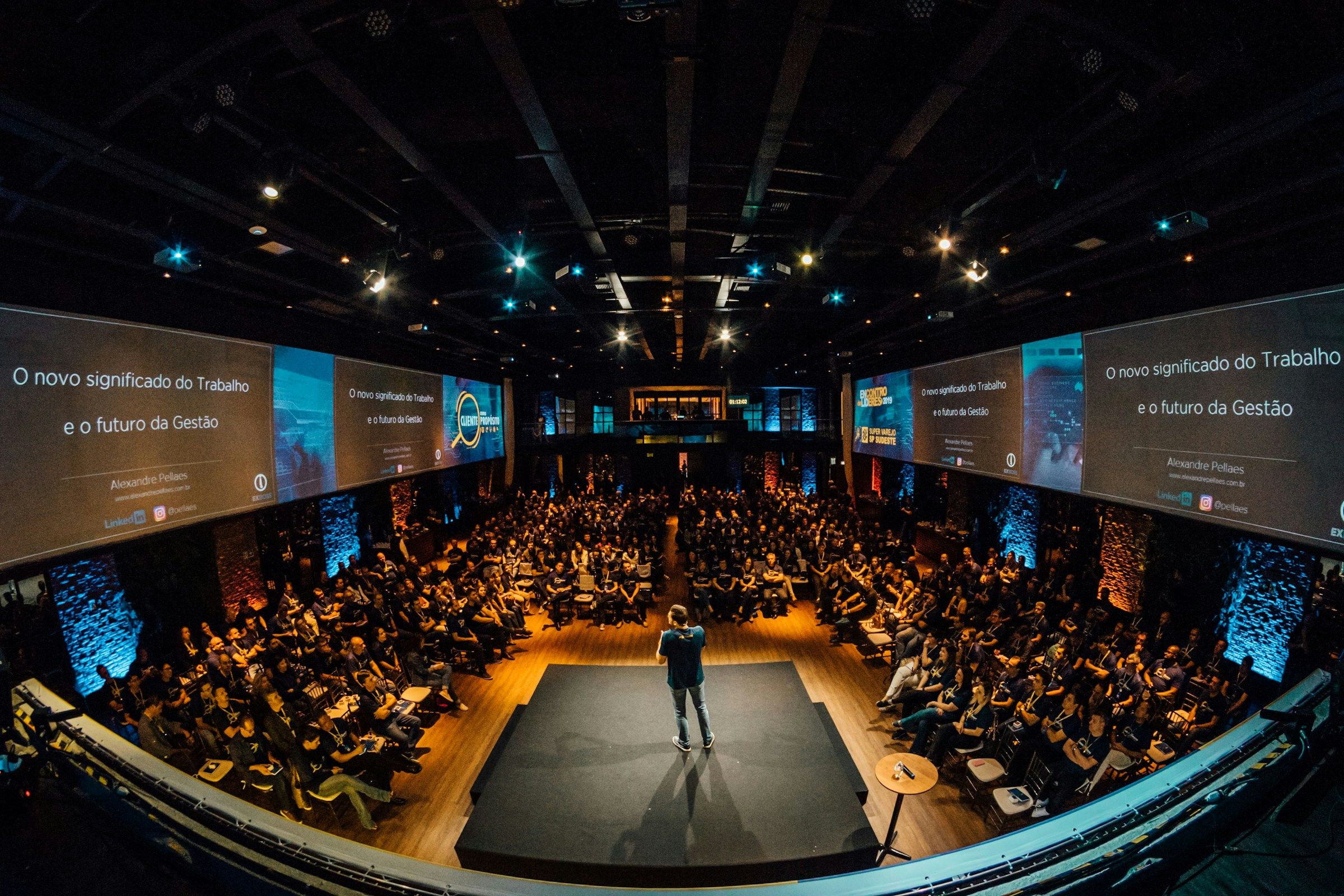 Auditorio lleno con un conferencista al centro y pantallas LED a los lados